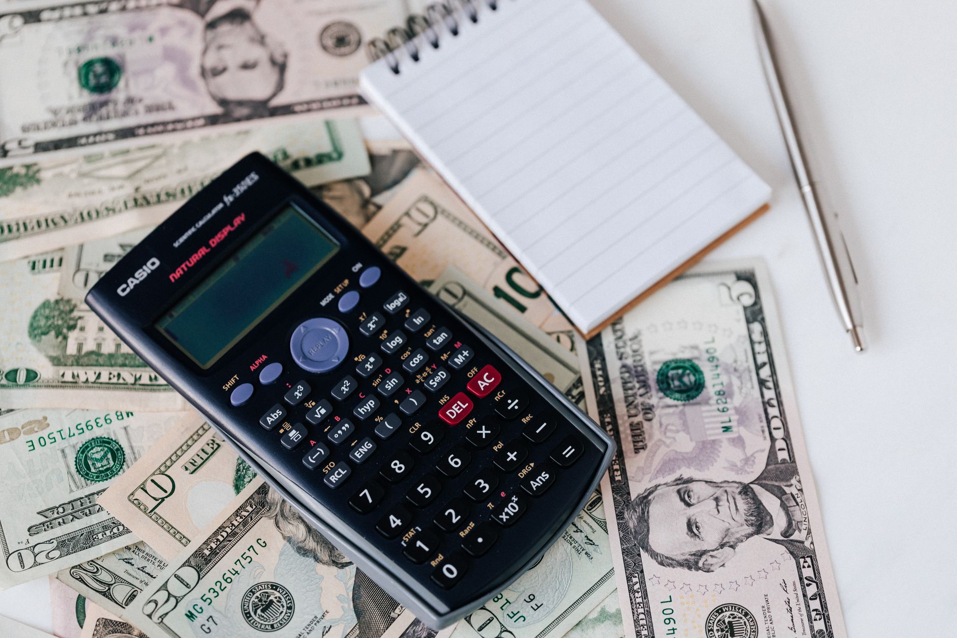 From above electronic calculator and notepad placed over United States dollar bills together with metallic pen for budget planning and calcu