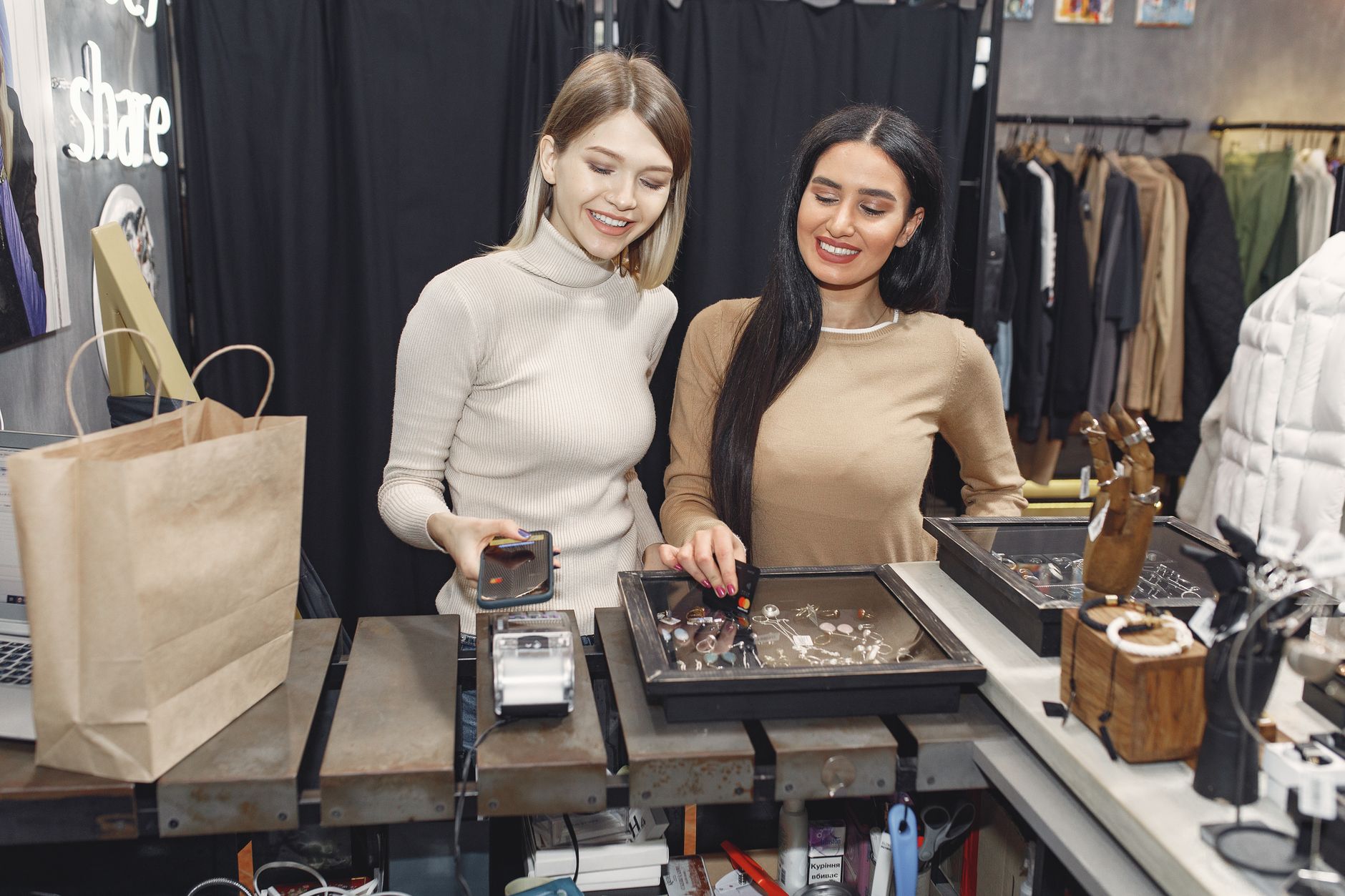 Cheerful casually dressed women paying with smartphone and credit card for purchases while standing near contactless terminal at counter des