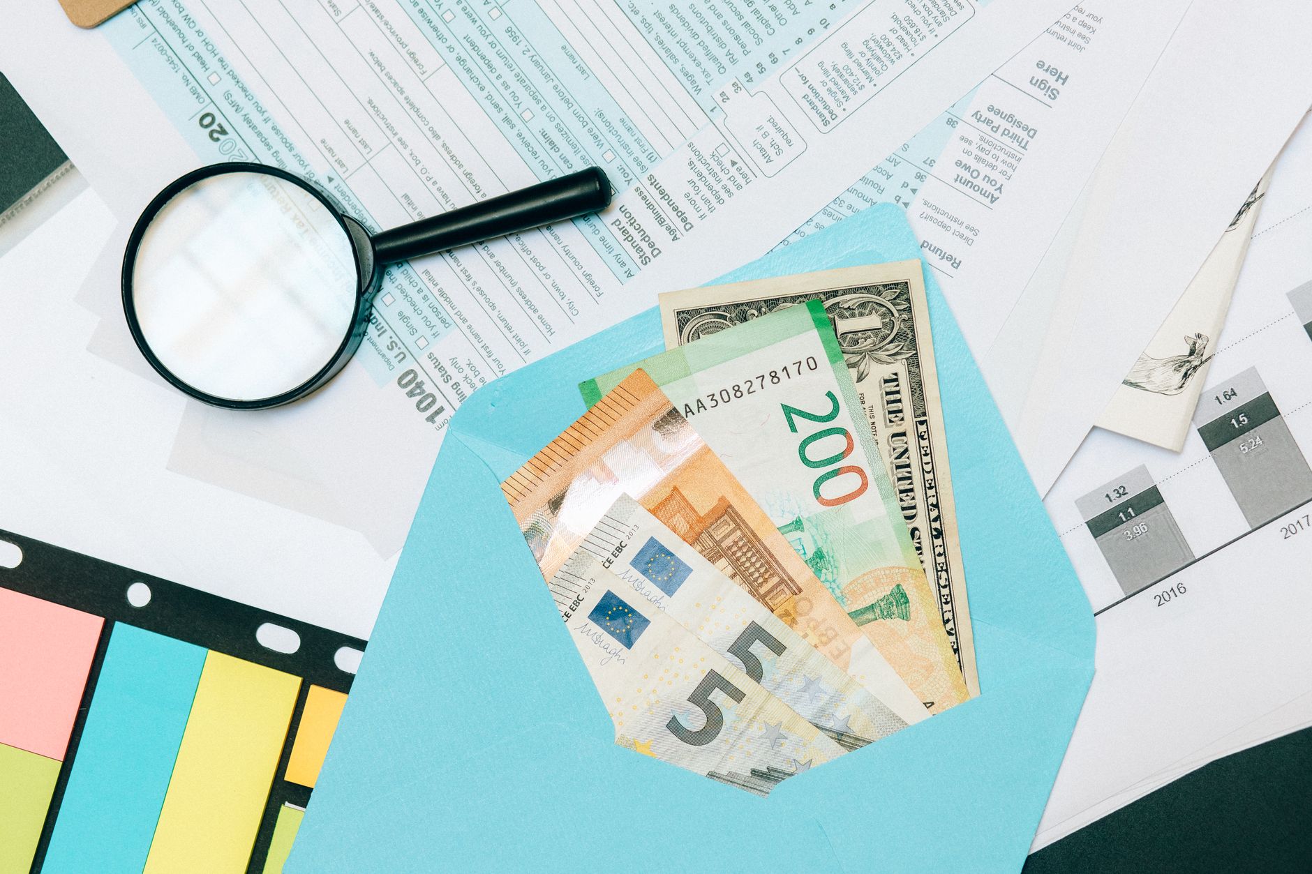 Close-up of tax documents and mixed currency in a blue envelope with a magnifying glass, perfect for financial analysis concepts.