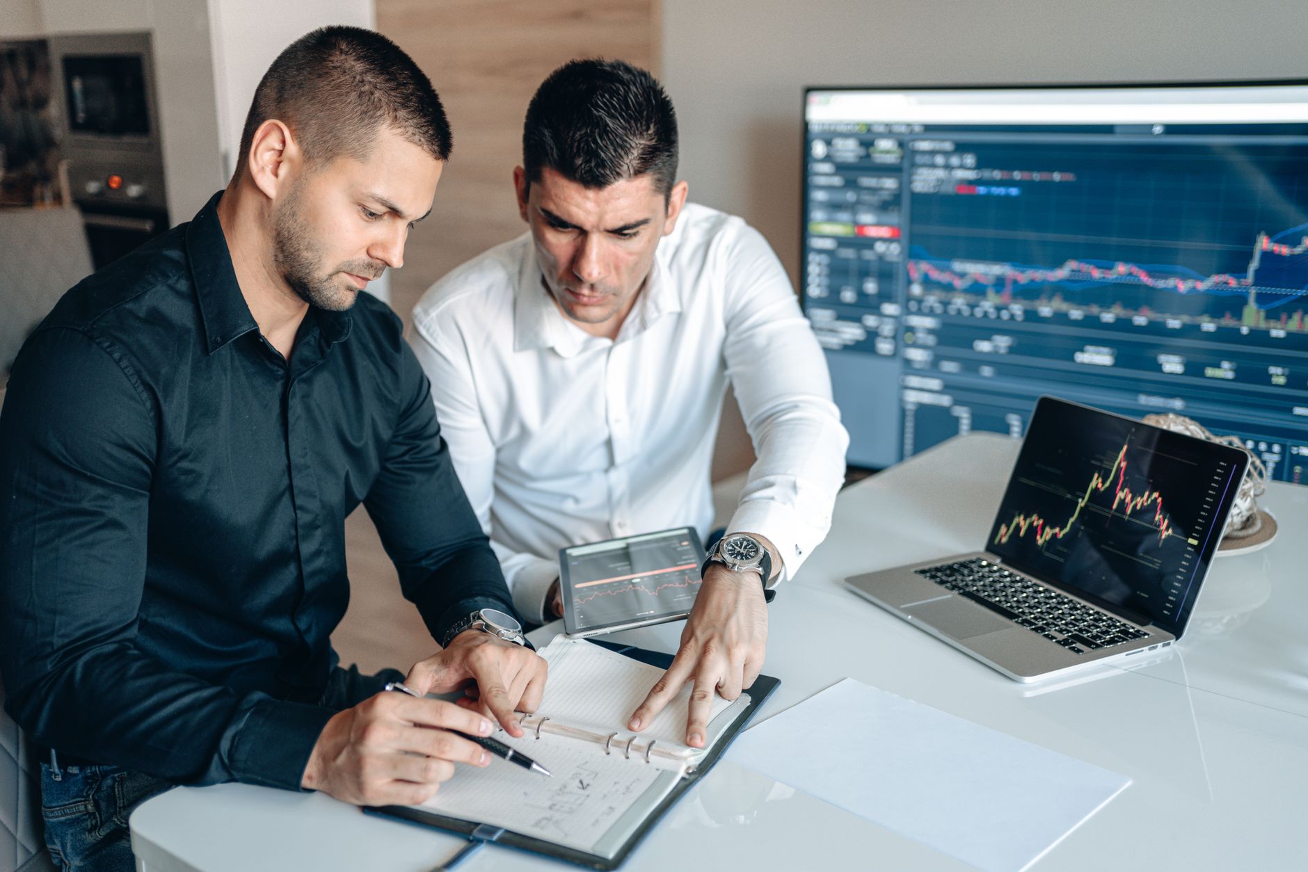 Two businessmen analyzing financial charts on digital devices in a modern office setting.